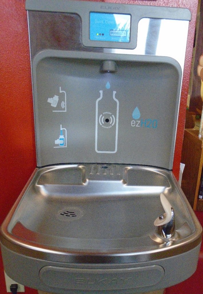 A stainless steel water fountain with a bottle of liquid on the side.